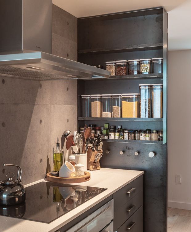 Compact contemporary kitchen with a dark open pantry and shelving unit, stocked with clear ingredient jars, spices, utensils, and cookware. The kitchen features a pale stone benchtop, dark cabinetry, black induction cooktop, stainless-steel rangehood, and grey concrete-look wall finishes. Warm task lighting highlights the practical storage and preparation area.
