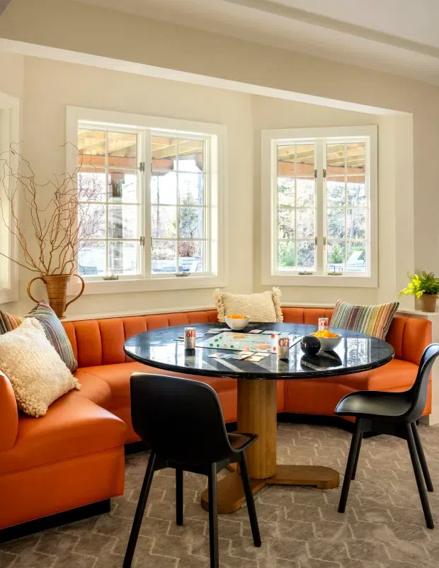 Bright, warmly styled dining nook with a curved burnt-orange upholstered banquette wrapping around a round black glossy dining table, paired with black dining chairs. The room has cream walls, white-trimmed multi-pane windows, exposed ceiling beams, patterned neutral carpet, decorative cushions, and a vase of branches. Sunlight enters through the windows, creating an inviting and colourful interior.