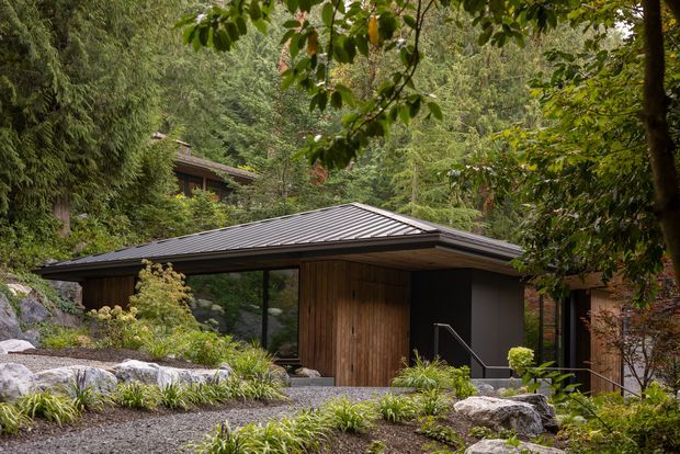 A low-profile contemporary pavilion-style dwelling nestled into a densely wooded landscape. The building features a broad dark metal hipped roof with deep overhangs, warm vertical timber wall cladding, large dark-framed glazing, and a minimalist dark entry volume. Natural boulders, gravel, and layered planting integrate the architecture into the forested garden setting.