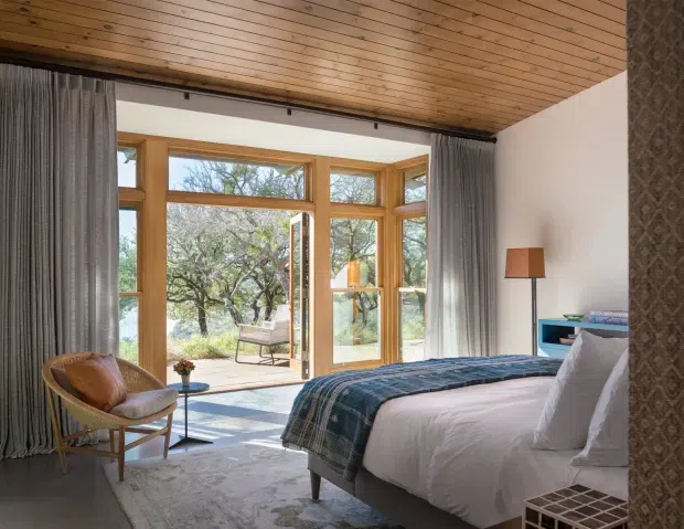 A warm, refined bedroom with a low timber-lined ceiling, a large timber-framed bay of floor-to-ceiling glazing and glazed doors opening toward a garden. The room contains a partially visible upholstered bed with white bedding and a blue patterned throw, a woven lounge chair, side table, floor lamp, curtains, rug, and a small teal cabinet. Natural light and views of trees create an inviting indoor-outdoor atmosphere.