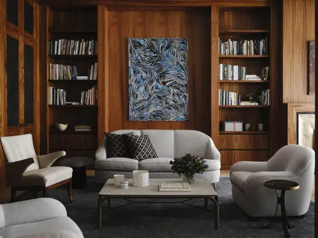 A sophisticated living room with dark timber wall panelling and integrated bookshelves surrounding a central abstract artwork. The room features a grey sofa, sculptural upholstered armchairs, a low rectangular coffee table, dark carpet, patterned cushions, and a fireplace edge at right. The overall composition is warm, refined, and library-like with strong mid-century influences.