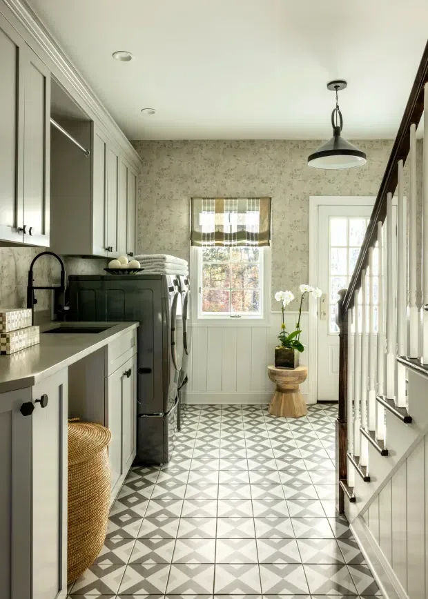 A narrow, bright laundry and utility room with pale built-in cabinetry, a stone-look benchtop, dark appliances, patterned geometric floor tiles, a farmhouse-style sink, stacked laundry appliances, and a staircase with white balustrade. Floral wallpaper, a window, glazed door, pendant light, and woven basket add traditional decorative character.