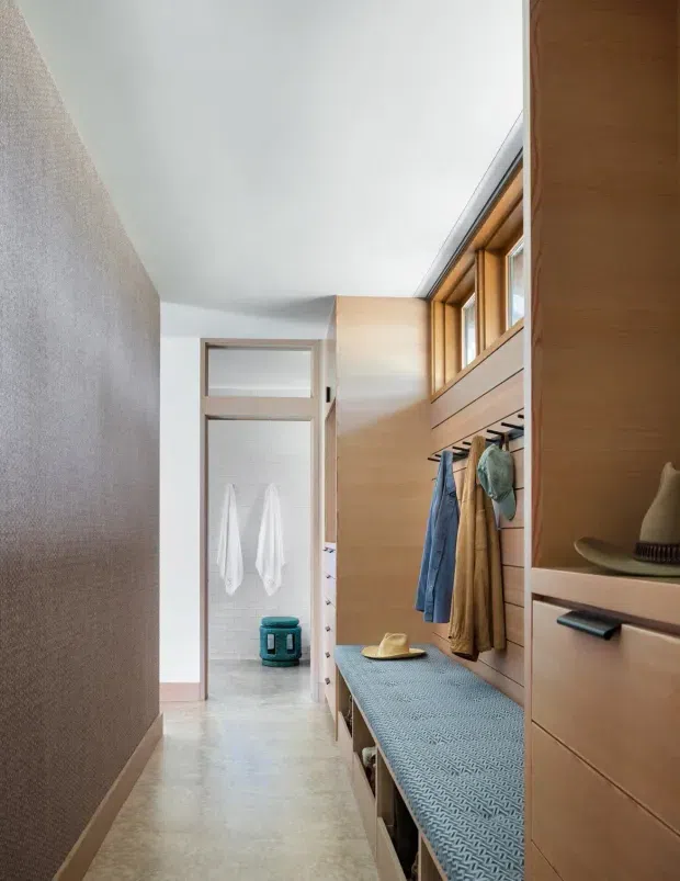 A bright, narrow dressing and entry corridor with extensive built-in timber cabinetry, an upholstered bench seat, open hanging storage, and high clerestory-style windows. A bathroom with white tiled walls is visible at the end of the passage. The space has a clean, warm modern appearance with pale flooring and natural wood joinery.