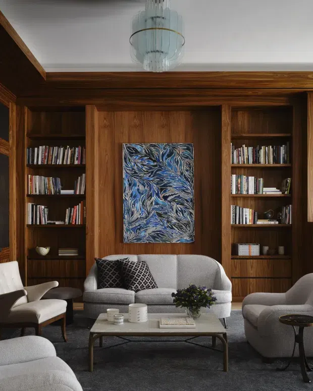 A refined living room with extensive dark timber built-in bookcases and panelled cabinetry surrounding a central artwork. The room includes a curved grey sofa, upholstered armchairs, a low rectangular coffee table, patterned cushions, and a sculptural glass pendant light. The palette is warm timber, charcoal grey, soft grey, and blue accents, creating a sophisticated, cosy atmosphere.