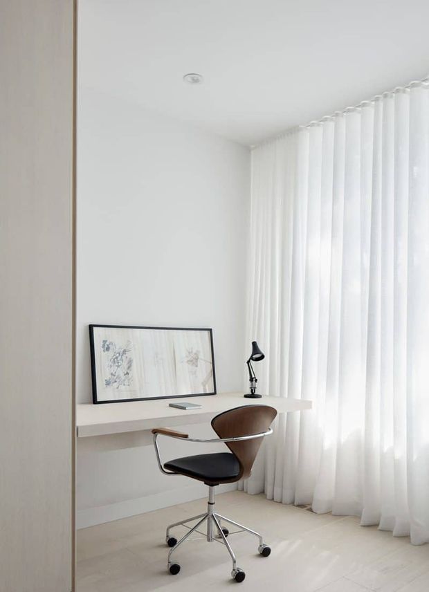 Bright, minimalist home office with a floating white desk positioned beside full-height sheer curtains. A black task lamp, framed artwork, and modern swivel chair create a restrained, uncluttered workspace. Pale walls and flooring produce an airy monochromatic interior.