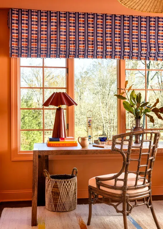 A richly coloured home office or study nook with terracotta-orange walls, tall timber-framed multi-pane windows, and patterned striped fabric blinds. A rustic wooden desk sits beneath the windows with a red table lamp, books and small decorative objects, accompanied by a woven basket and a bamboo or rattan framed chair. A potted plant adds greenery, while a pale rug with subtle coloured accents covers the floor. The room has a warm, eclectic, vintage-inspired character.