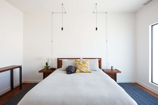 Bright, minimalist bedroom with a neatly made bed centered against a white wall. The bed has a dark timber headboard, white bedding, grey and yellow geometric accent cushions, and small timber bedside tables. Slim black pendant lights hang symmetrically on either side of the bed, while a large window provides strong natural light. A dark striped rug and timber flooring add contrast.