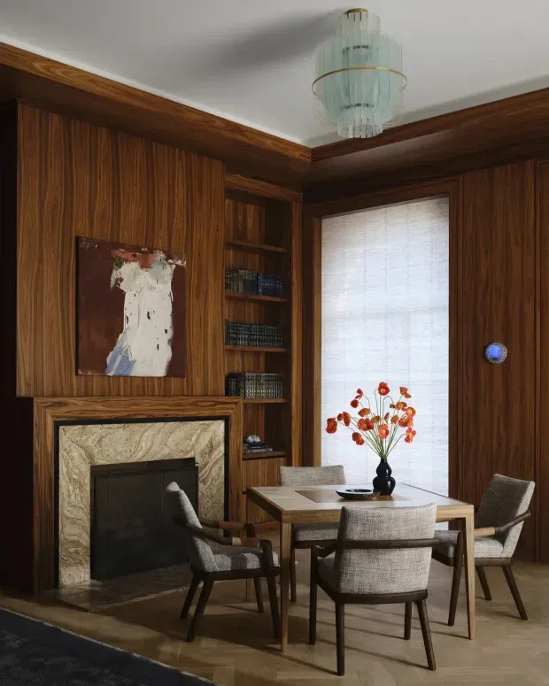 A richly paneled dining room with dark timber walls and built-in shelving, centered on a rectangular dining table surrounded by upholstered chairs. A marble-faced fireplace, artwork, books, a tall window with a woven blind, and a sculptural glass pendant light complete the warm, refined interior.