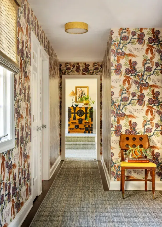 A narrow residential hallway lined with bold floral wallpaper, white trim and doors, a window with woven blind, and a grey patterned carpet runner. A small vintage wooden chair and side table create a decorative vignette, while a framed artwork and console are visible through the doorway at the end. The space is bright, richly patterned, and eclectic with warm timber and brass accents.