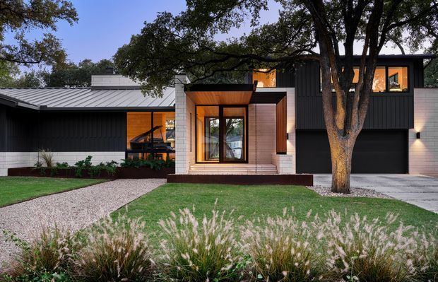 Contemporary single-storey residential facade with a low-pitched metal roof, dark vertical cladding, pale masonry, and a timber-lined recessed entry. Large mature trees frame the front elevation, with a gravel path, lawn, and landscaped ornamental grasses leading to the entrance. A dark garage door and warm-lit windows reinforce the modern, restrained composition.