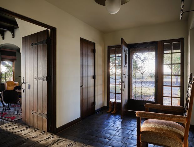 A warm, character-filled entry hall with dark timber trim, rustic plank and slate-like tiled flooring, and a glazed front door with decorative wrought-iron detailing. A substantial vertical-plank timber door opens toward an adjoining room, while a pair of woven-seat chairs sits near the entry. The space has traditional architectural detailing and abundant natural light.
