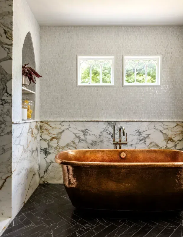 A refined bathroom featuring a freestanding copper-toned bathtub, dark herringbone tile flooring, dramatic veined marble wall cladding, and a small tiled niche with open shelving. The upper walls are finished in a light textured mosaic tile, with two white-framed arched windows bringing in natural light and views of greenery.