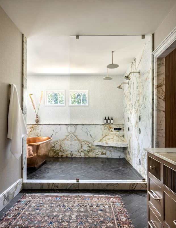 A luxurious bathroom with a large walk-in shower enclosed by clear glass, featuring dramatic veined marble walls, a marble shower bench and threshold, dark herringbone floor tiles, a copper freestanding bathtub, brass-toned fixtures, and a timber vanity. A patterned vintage-style rug and white towels add warmth and texture.