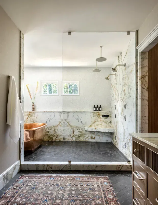 Luxurious bathroom viewed from the vanity area, featuring a walk-in marble-clad shower with multiple fixtures, a copper freestanding bathtub, dark herringbone tile flooring, patterned rug, and warm wood cabinetry. A frameless glass shower partition and arched windows contribute to the refined, light-filled interior.