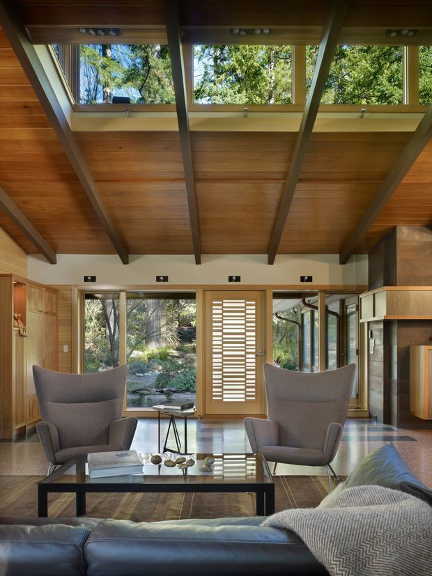 A warm, wood-lined living room with a dramatic exposed-beam ceiling and clerestory glazing overlooking a wooded landscape. The room features two sculptural high-back lounge chairs, a dark sectional sofa, a glass-topped coffee table, timber wall panelling and cabinetry, polished flooring, and a distinctive slatted timber entry door. Abundant glazing and skylight-like clerestory windows create a strong connection to nature.