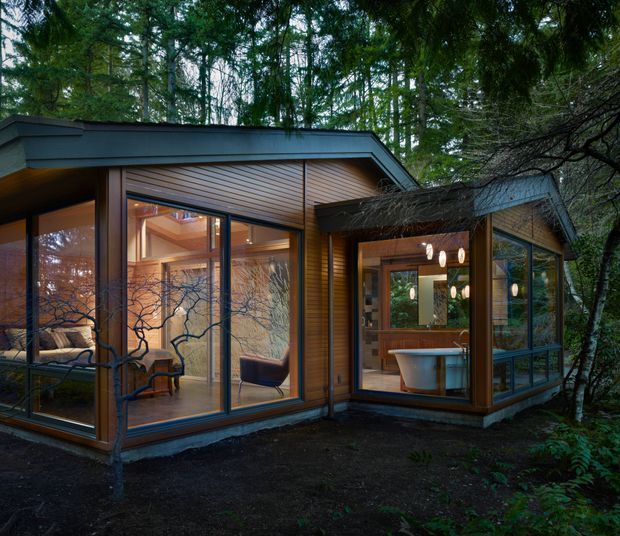 Exterior view of a compact modern cabin or house set among dense woodland. The building has warm timber cladding and extensive floor-to-ceiling glazing, with a low-pitched intersecting roof form. Through the windows, a furnished living area and a bathroom with a freestanding tub are visible. The dark forest setting and warm interior lighting create an intimate, secluded atmosphere.