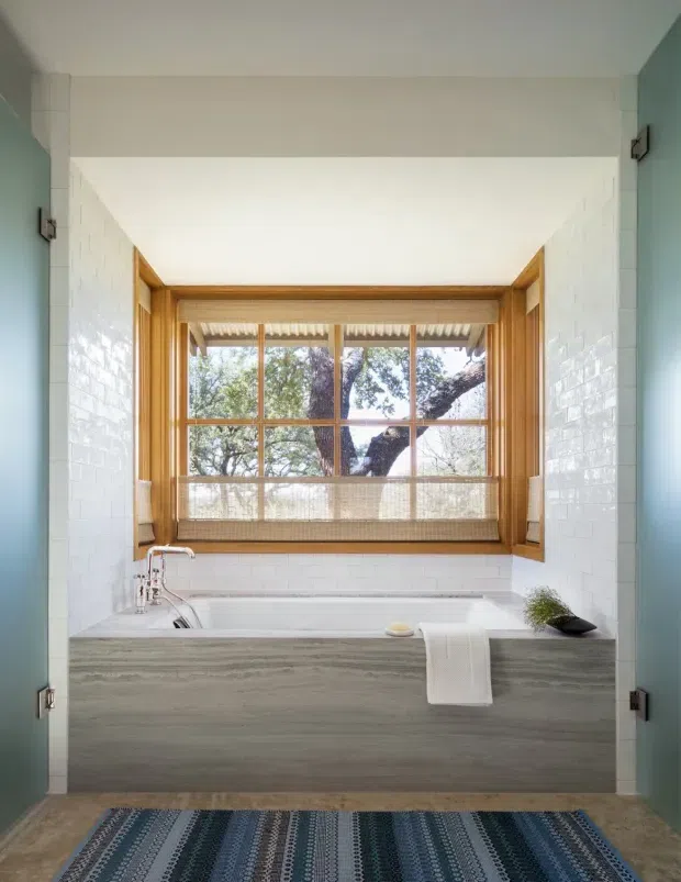 A bright bathroom centred on a built-in bathtub beneath a large timber-framed window. The tub surround has a grey wood-grain finish, while glossy white subway tiles line the side walls and window recess. Pale blue-green doors or wall panels frame the opening, with polished chrome tapware, a woven blue patterned rug, and a small plant adding understated decorative detail. The window overlooks leafy trees and brings abundant natural light into the space.