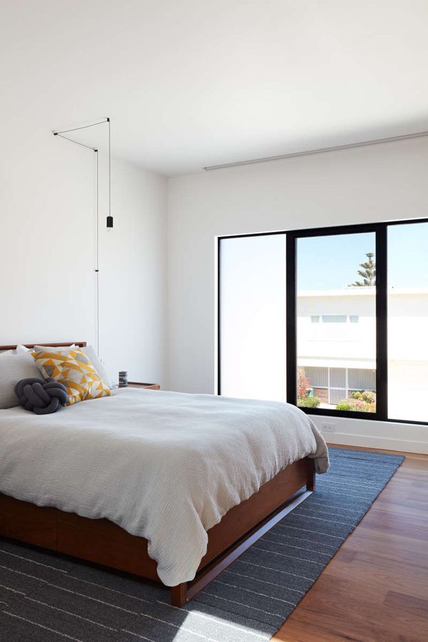 Bright, minimalist bedroom with a low-profile timber bed dressed in a light quilt, patterned accent cushions, and a dark grey striped rug over timber flooring. Large black-framed windows provide abundant natural light and views of neighbouring architecture. A simple exposed hanging pendant and slim linear ceiling fixture reinforce the clean contemporary aesthetic.