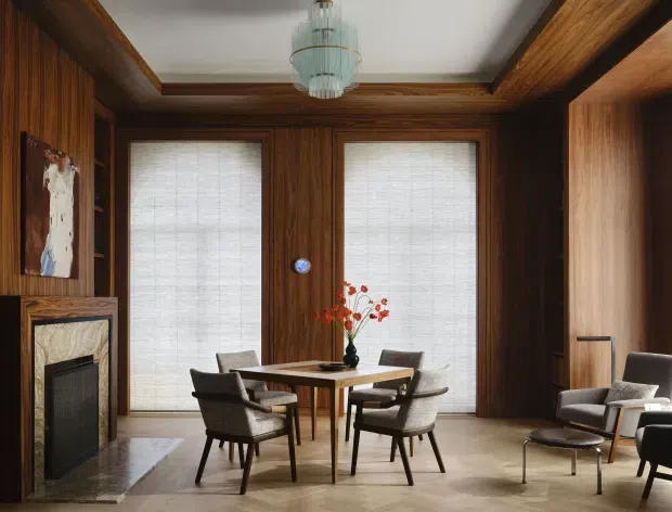 Elegant dining room with rich dark wood wall panelling, matching ceiling beams and built-in cabinetry, a central rectangular dining table surrounded by upholstered chairs, a marble fireplace surround at left, tall screened windows, and a sculptural pendant light. The room has a refined, warm, mid-century-inspired character with strong timber detailing.