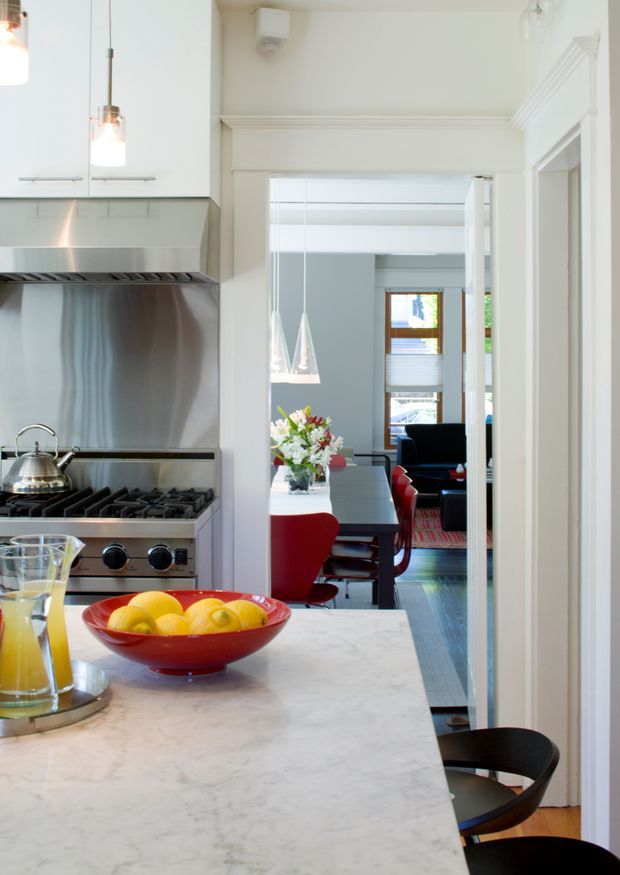 A bright, narrow kitchen opening toward a dining and living area. The kitchen features white cabinetry, a large pale stone island or benchtop, stainless-steel splashback and rangehood, gas cooktop, pendant lighting, and dark timber flooring. Through the wide opening, a dining table with red chairs and a lounge area are visible, with traditional moulded door and ceiling trims throughout.