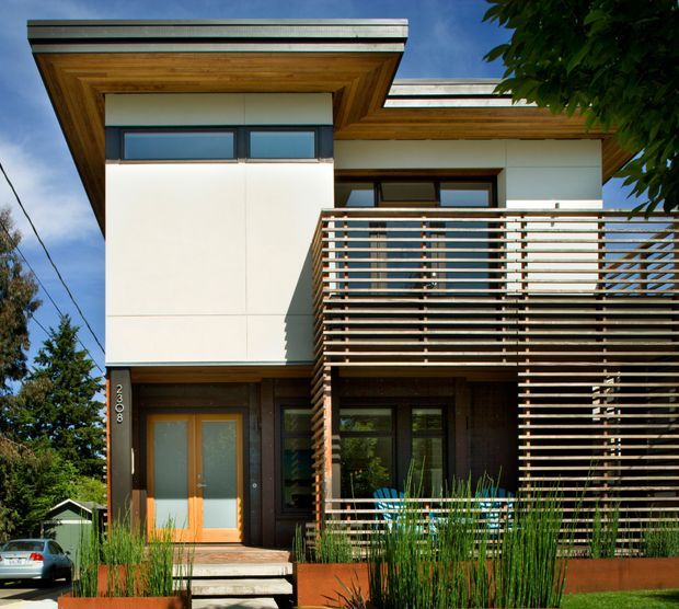 Exterior view of a contemporary multi-storey residence with strong horizontal lines, projecting flat roof forms, white rendered wall panels, dark window and door frames, warm timber soffits, and a prominent slatted timber privacy screen and balcony balustrade. The front entry is approached by steps and surrounded by designed planting.