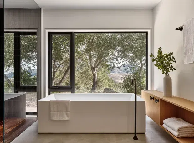 Contemporary bathroom featuring a freestanding white bathtub positioned in front of large black-framed windows, with a freestanding dark tap and a timber vanity or storage bench with open towel shelving. Neutral plaster walls, a smooth grey floor, and warm timber flooring visible through the adjacent opening create a refined, natural setting. Trees and mountainous terrain are visible beyond the glazing.