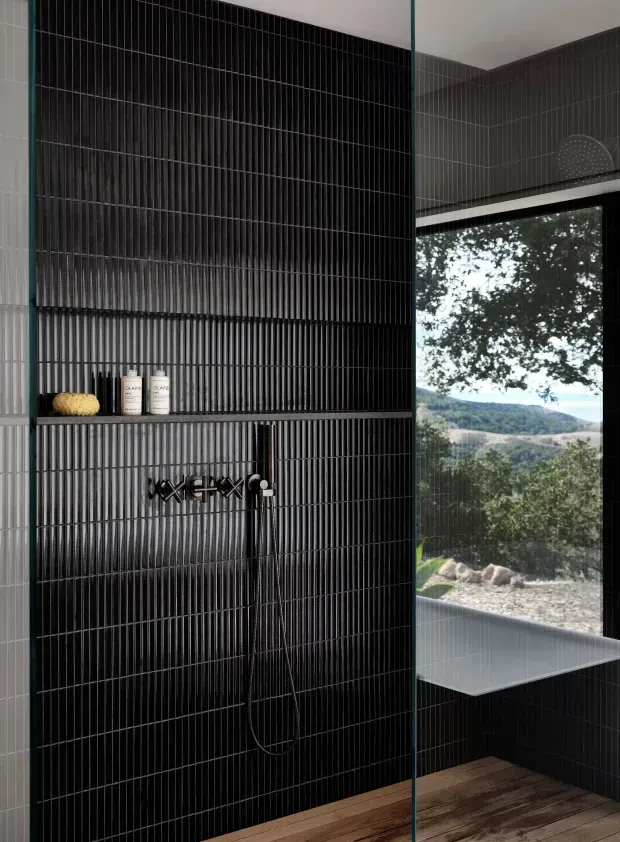 Contemporary bathroom shower with a dramatic black vertically ridged tile feature wall, integrated recessed shelf, and dark wall-mounted shower fittings. A frameless glass screen separates the shower from a deep grey tiled area with a built-in window seat or ledge, opening to a leafy landscape view. The floor appears to be dark timber or timber-look material, creating contrast with the black tilework.