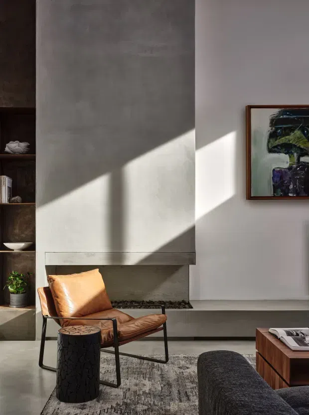 A refined contemporary living room with a tall minimalist fireplace clad in textured grey plaster or concrete, dramatic sunlight casting geometric shadows across the walls, dark built-in shelving, a caramel leather lounge chair, textured rug, dark upholstered sofa, and timber coffee table. Abstract artwork and restrained furnishings create a warm, gallery-like atmosphere.