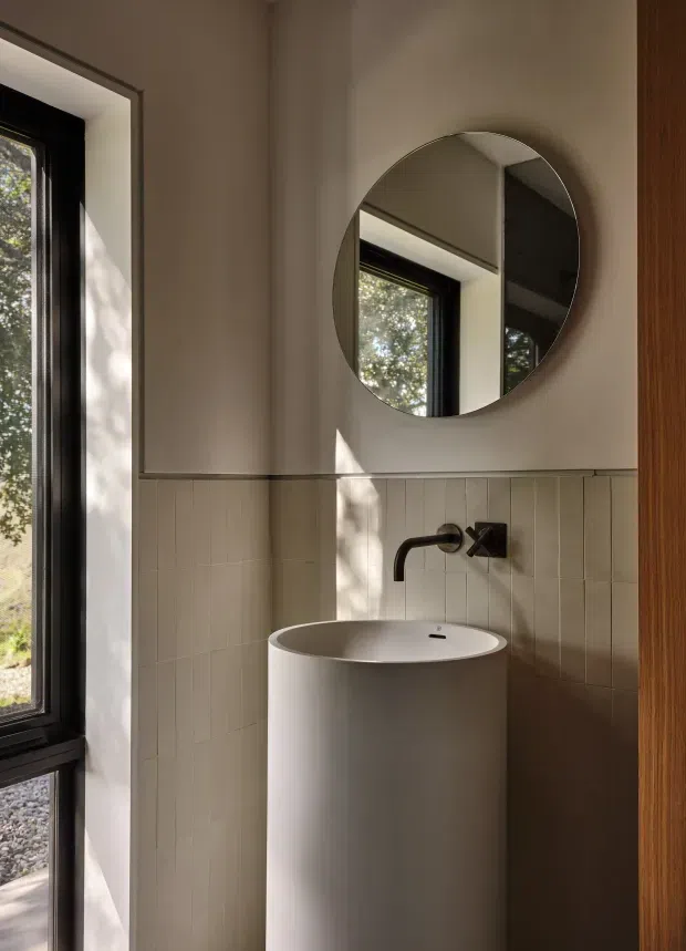 A compact, refined bathroom wash area featuring a freestanding cylindrical pedestal basin, wall-mounted dark metal tapware, a round frameless mirror, and light vertical rectangular wall tiles beneath smooth warm beige painted walls. A black-framed window provides natural light, while a timber-toned door or joinery edge is visible at right.