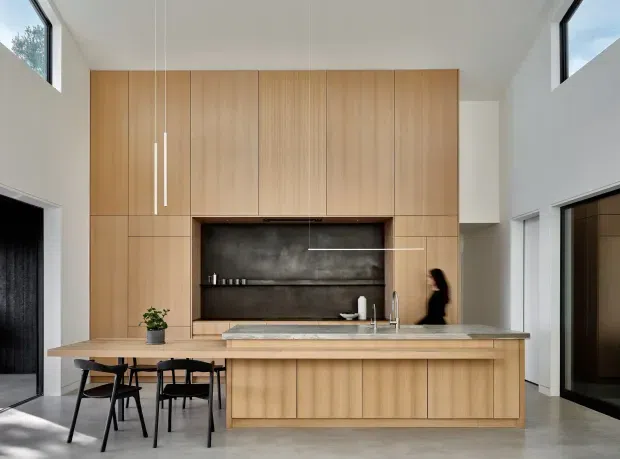 A contemporary open-plan kitchen dominated by a full-height light timber cabinetry wall with an integrated dark stone or concrete-look cooking and display recess. A matching timber island with a pale stone benchtop includes a sink and tap, while a small dining setting sits at one end. The room has white walls, a high ceiling, clerestory-style windows, minimalist pendant lights, and dark-framed glazed openings.