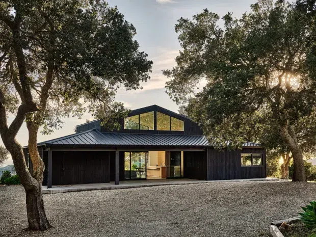 A low, modern dark-clad house set among mature trees, viewed from a broad gravel forecourt. The building has a charcoal standing-seam metal gable roof, black vertical cladding, large glazed openings, a prominent raised central gable with clerestory-style glazing, and an integrated garage at the left. Warm interior light is visible through the front glazing.