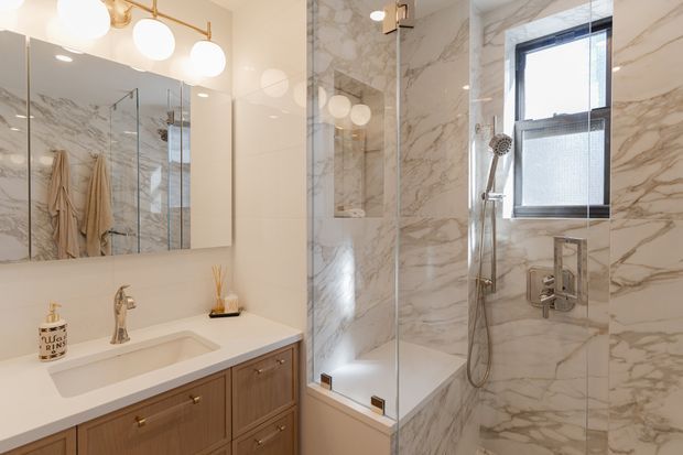 A bright, refined bathroom featuring a floating-style timber vanity with a white integrated basin, brushed metal tapware, large wall mirrors, and a glass-enclosed shower over a bath. The walls and shower surround are clad in dramatic white and grey veined marble-look tile, complemented by globe vanity lighting and warm brass-toned hardware. A window provides natural light.