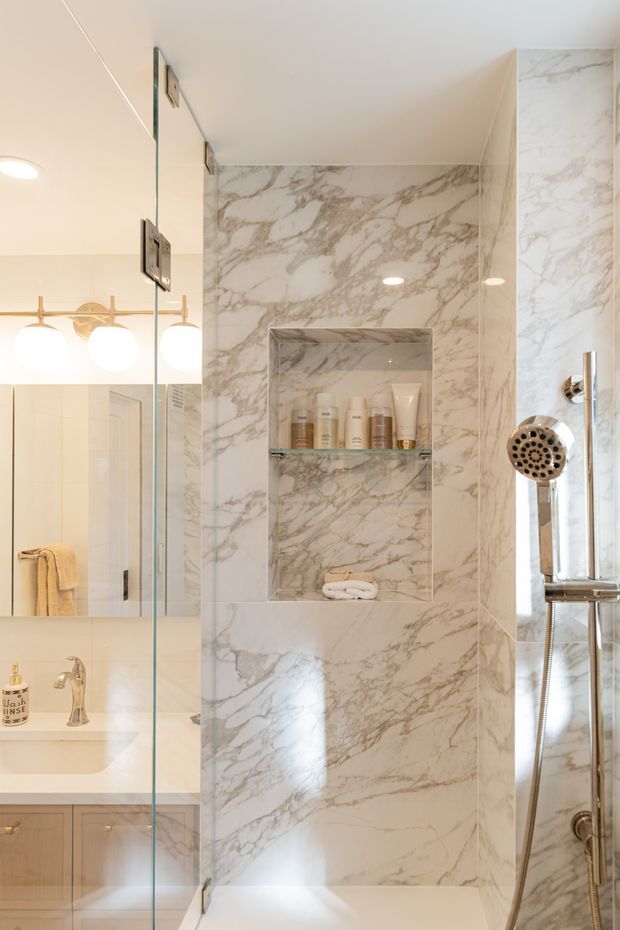 Bright, refined bathroom focused on a glass-enclosed shower clad in white marble-look veined tile. The shower includes a recessed niche with toiletries and folded towels, chrome fixtures and a handheld shower. A mirrored vanity with a pale stone countertop, light wood cabinetry and globe wall lights is visible to the left.