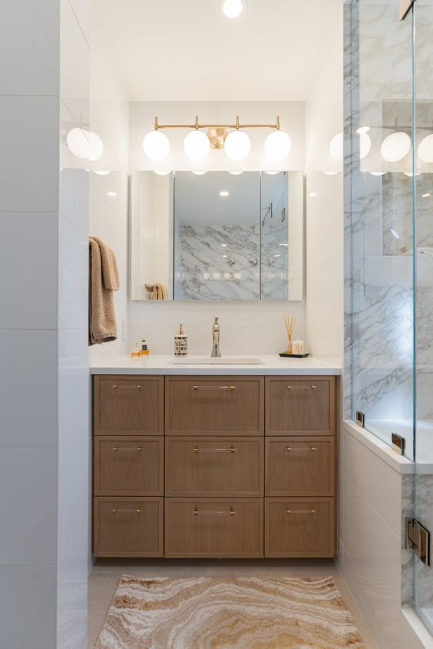 Bright, compact contemporary bathroom vanity area with a large mirrored wall, marble-look tiled surfaces, a white benchtop and integrated basin, and extensive warm wood cabinetry. Brass-toned hardware and a multi-globe vanity light add a refined accent, while a glass-enclosed shower or bath area is visible on the right.