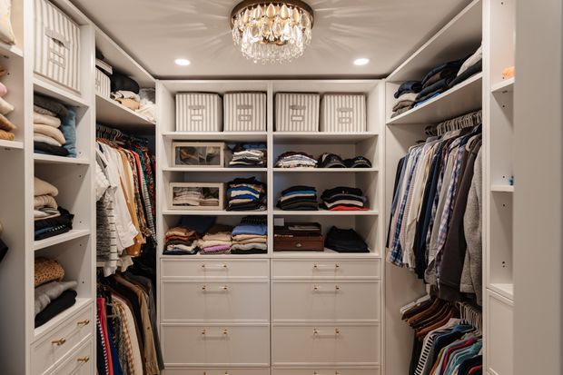 A compact, highly organized walk-in wardrobe with white built-in cabinetry, open shelving, drawers, cubby storage, and hanging rails filled with folded and hanging clothing. A decorative crystal chandelier and recessed ceiling lights provide warm, refined illumination.