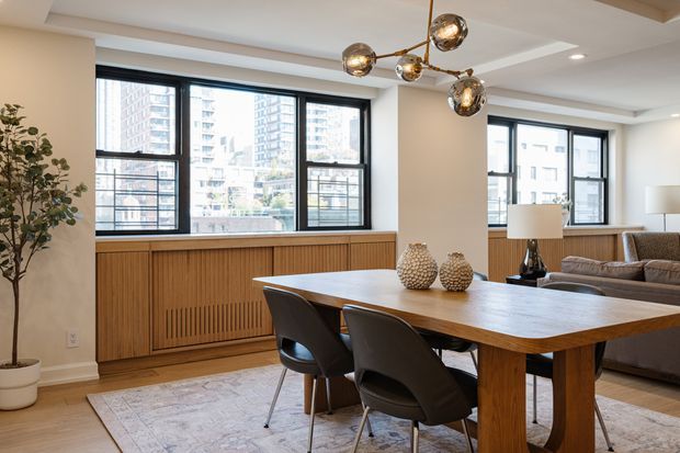 Bright contemporary open-plan dining and living area in an urban apartment, featuring large black-framed windows, a warm timber dining table and slatted timber window bench or radiator cover, neutral walls, mid-century-inspired dining chairs, a grey sofa, patterned area rug, and sculptural spherical pendant lighting. High-rise buildings are visible outside.