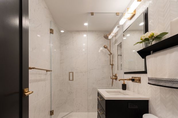 A compact, refined bathroom viewed from the doorway, featuring a glass-enclosed shower with marble-look wall and floor tiles, a white vanity basin with dark cabinetry, a large black-framed mirror, and brushed brass or bronze fixtures. A patterned towel and small floral arrangement add decorative detail. Recessed lighting and a bright neutral palette create a polished contemporary feel.