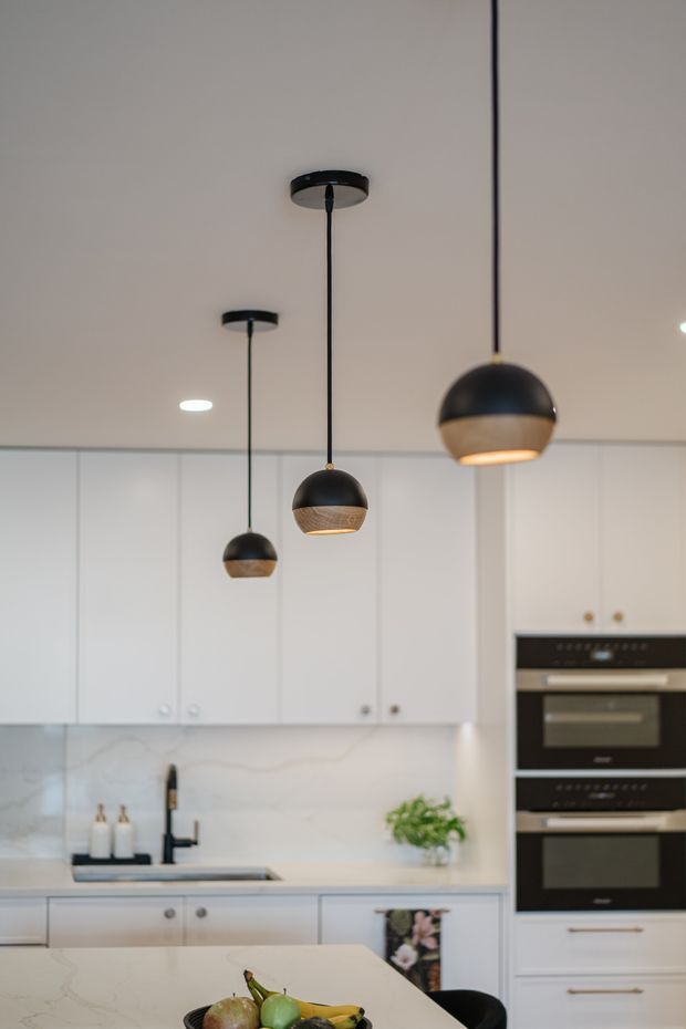 Bright contemporary kitchen with white flat-panel cabinetry, a pale veined stone splashback and benchtops, integrated black double ovens, and a large island. Three black pendant lights with timber-toned lower shades hang over the island, creating a warm focal detail. A sink, black tap, small plant, and fruit bowl are visible.