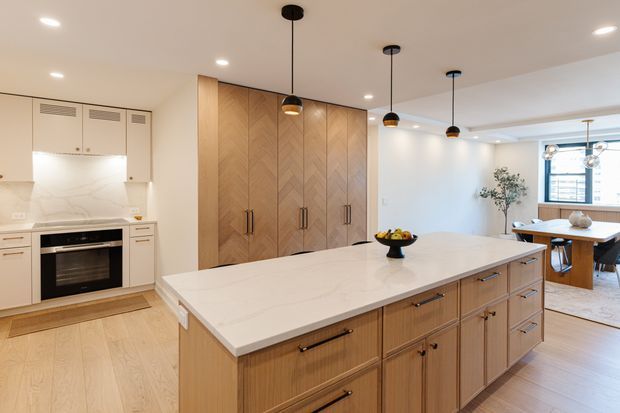 Bright open-plan kitchen dominated by a large island with a pale stone benchtop and light timber cabinetry. The kitchen includes integrated appliances, a full-height chevron-patterned timber pantry or storage wall, pendant lighting, recessed ceiling lights, and an adjacent dining area visible beyond. The space has a warm, refined contemporary appearance with generous circulation and natural light.