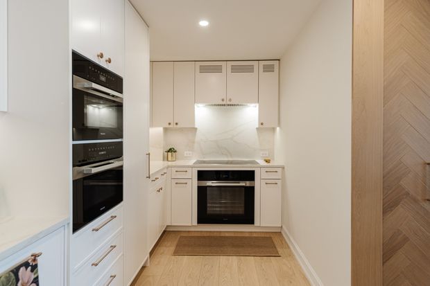 A compact, refined kitchen with a U-shaped arrangement of white painted cabinetry, integrated black ovens, a dark cooktop and oven, pale stone-look splashback, and light timber flooring. Brass or warm metal handles add subtle contrast. A timber chevron-patterned door or wall panel is visible at the right edge, and a small mat sits in front of the cabinetry. The space is bright, clean, and highly tailored, with built-in ventilation grilles above the cooking area.