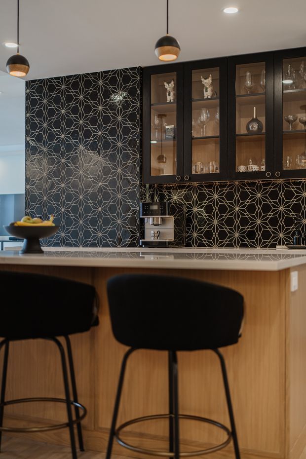 A stylish kitchen and bar area featuring a timber island with a pale stone benchtop, two black upholstered bar stools, and black-framed glass-front cabinetry displaying glassware. A bold black geometric patterned splashback and matching feature wall create a strong decorative focal point. Black-and-brass pendant lights and warm timber finishes add a refined, dramatic atmosphere.