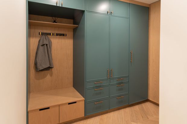 Built-in entry storage and wardrobe joinery with muted teal-blue cabinetry, brass-toned handles, and warm timber interiors. The unit includes tall cupboards, drawers, an open coat-hanging niche, and a timber bench with lower storage. Light timber flooring and warm recessed lighting create a refined, functional entrance storage area.
