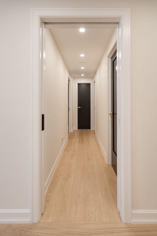 A narrow, clean residential hallway viewed through a trimmed doorway. The corridor has pale painted walls and ceiling, recessed downlights, light natural timber flooring, white skirting boards and door casings, and several internal doors including a dark charcoal door at the far end. The space is uncluttered and softly lit with a contemporary, neutral appearance.