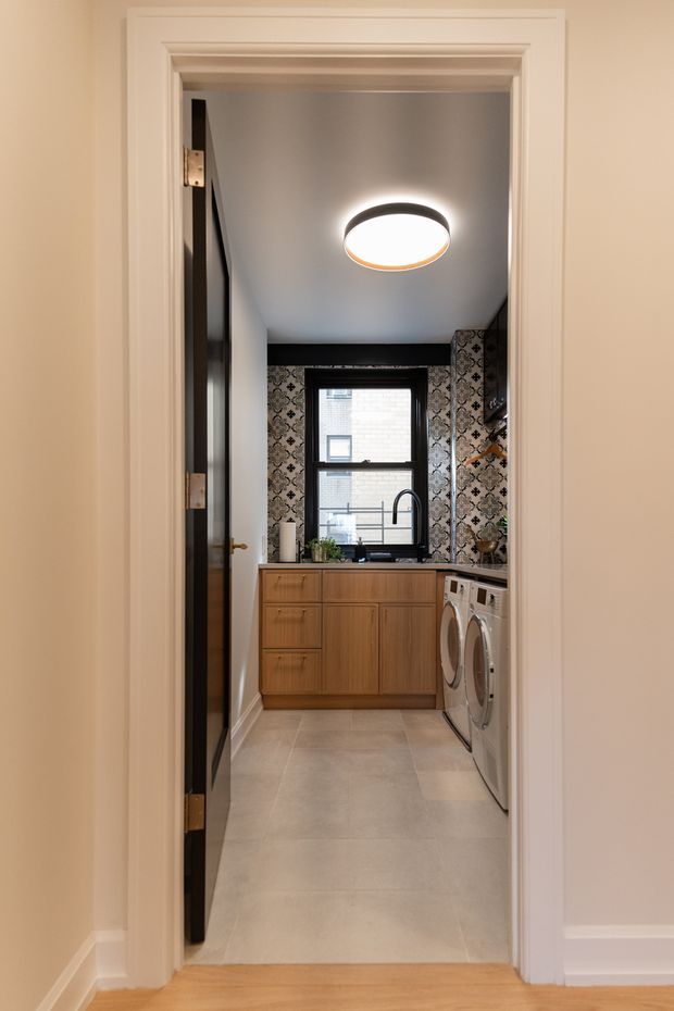 View through a trimmed doorway into a compact, well-designed laundry room. The room features warm timber cabinetry, a pale large-format tiled floor, patterned dark decorative wall tiles around a black-framed window, a black tap, front-loading washing machines, and a round flush-mount ceiling light. White painted door trim and warm timber flooring in the foreground create a refined transitional feel.