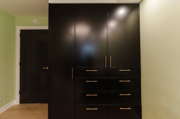 A compact bedroom or dressing area dominated by a floor-to-ceiling dark built-in wardrobe and chest of drawers with slim brass-toned handles. The room has pale green walls, a black interior door, light timber flooring, white trim, and simple recessed ceiling lighting.