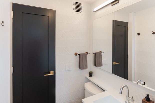 Bright contemporary bathroom vanity area with a large wall mirror, white textured geometric wall finish, white countertop and basin, black paneled door, brass hardware, towel hooks, and integrated linear ceiling lighting. The composition is focused on the bathroom interior, with the black door and mirror adding strong contrast.