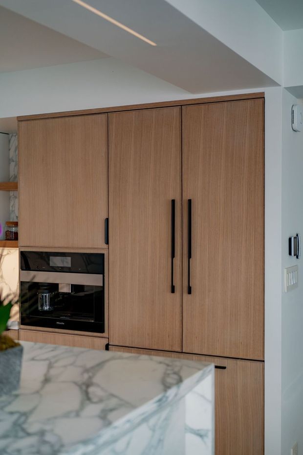 Close-up view of a contemporary kitchen featuring full-height warm wood-grain cabinetry with long black handles, integrated black-glass appliances, and a prominent white marble island with grey veining in the foreground. White walls and a recessed ceiling with linear lighting complete the clean, refined fit-out.