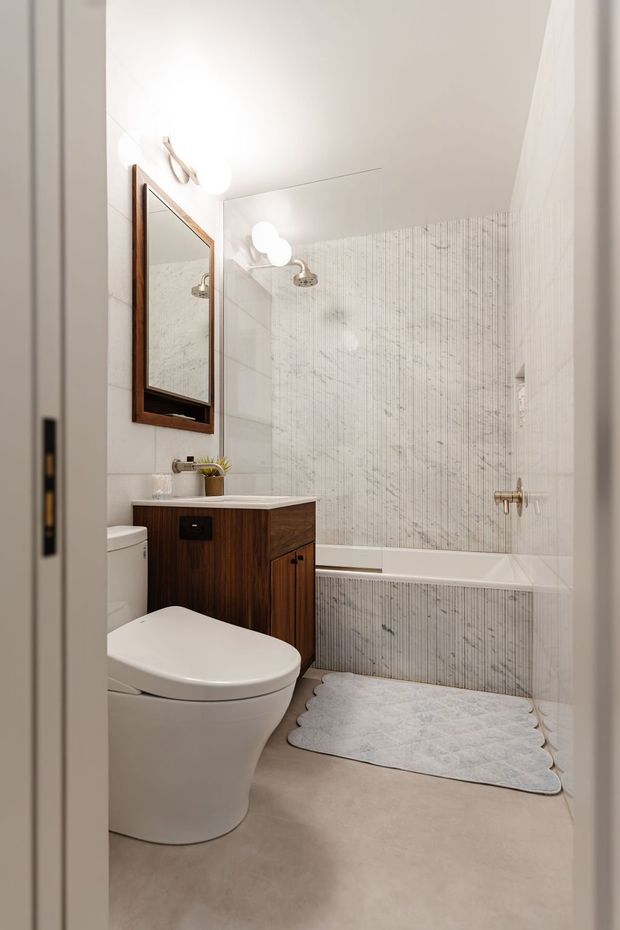 Compact contemporary bathroom viewed through the doorway, featuring a white toilet, dark timber vanity with a white benchtop, framed mirror, bathtub with shower, and pale vertically textured wall tiles. The room has warm neutral finishes, brass-toned fittings, and soft globe wall lighting.