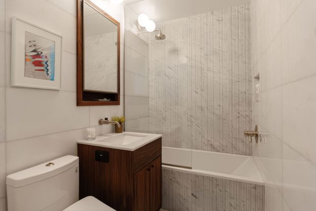 Compact contemporary bathroom with a white toilet, wall-mounted wood vanity, integrated white basin, framed mirror, and white bathtub enclosed by vertically textured light tile. Brass-toned tapware and simple globe lighting add warm accents.