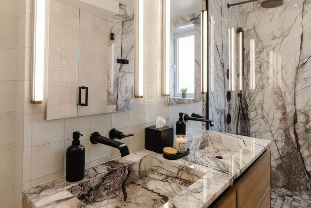 A refined bathroom vanity and shower area with a double vanity, black wall-mounted taps, white cabinetry, and dramatic veined marble surfaces. Large mirrors, vertical illuminated sconces, a frameless glass shower enclosure, and marble-clad walls create a luxurious contemporary interior.